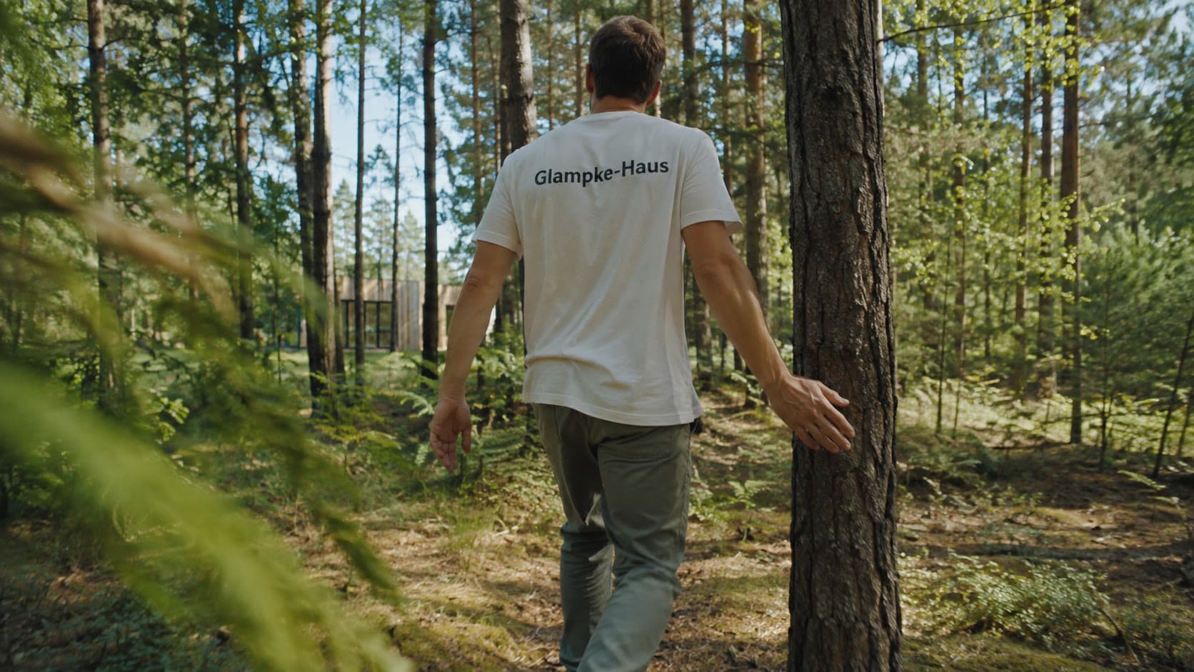 Ein Mitarbeiter von Glampke-Haus geht durch einen Kiefernwald und legt die Hand an einen Stamm, im Hintergrund ein Holzhaus
